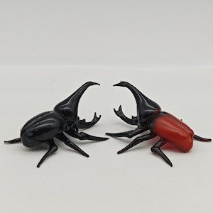 Könnte beinhalten: Zwei Glas-Käfer-Figuren, eine schwarze und eine rote, die sich gegenüberstehen. Die Käfer haben lange Hörner und detaillierte Beine.