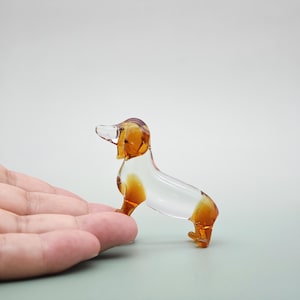 May include: A clear glass dachshund figurine with amber-colored accents on its head, ears, and legs. The small dog sculpture is posed in a standing position, showcasing its elongated body and short legs. The figurine is a decorative item.