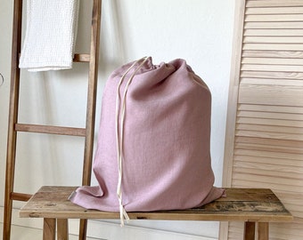 May include: A dusty pink linen laundry bag with a white drawstring sits on a wooden bench. The bench has a rustic, weathered look and is made of natural wood.