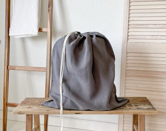 May include: A grey linen laundry bag sits on a wooden bench. The bag has a white drawstring and is made of a soft, natural fabric.