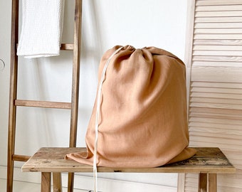 May include: A large, peach-coloured linen laundry bag with a drawstring closure sits on a rustic wooden bench. A wooden ladder with a white towel is to the left. The background includes a white wall and a slatted wooden screen.