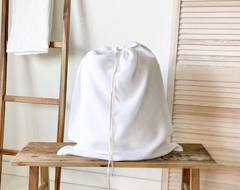 May include: A white laundry bag with a drawstring closure sits on a wooden bench. The bench has a rustic, weathered look and is made of natural wood.