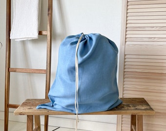 Puede incluir: Una bolsa de lavandería azul claro con un cordón de ajuste blanco se encuentra sobre un banco de madera. El banco tiene un aspecto rústico y desgastado.