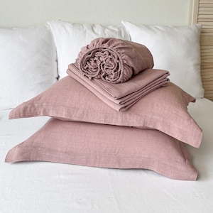 May include: A set of pink linen bedding, including a fitted sheet, flat sheet, and two pillowcases. The bedding is folded neatly on a white bed.