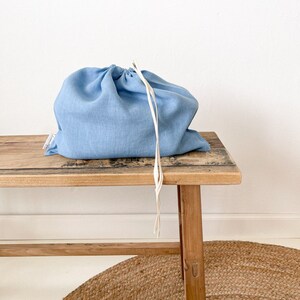 May include: A light blue linen drawstring bag sits on a wooden bench. The bag has a white drawstring and is made of a soft, natural fabric.