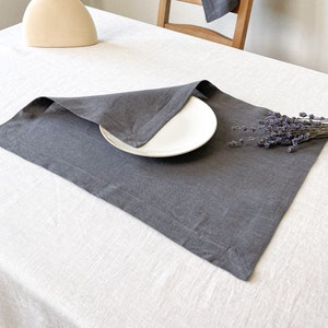 May include: A gray linen placemat on a white linen tablecloth. A white plate is on top of the placemat. A bouquet of dried lavender is on the table.