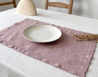 May include: A dusty rose linen placemat with a white plate on top. The placemat is on a white linen tablecloth. There is a vase with dried wheat stalks in the background.