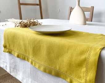 May include: A yellow linen table runner with a stitched border sits on a white tablecloth. A white ceramic vase with a dried wheat stalk sits on the table.