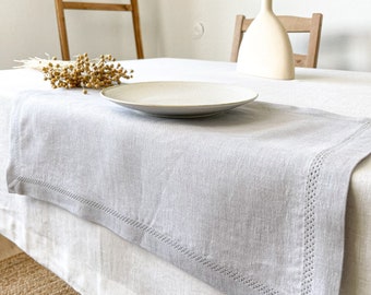 Puede incluir: Un camino de mesa de lino gris claro con un borde cosido se encuentra sobre un mantel blanco. Un jarrón de cerámica blanco con una espiga de trigo seca se encuentra sobre la mesa. Un plato blanco se encuentra sobre el camino de mesa.