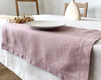 May include: A dusty rose linen table runner with a stitched border sits on a white tablecloth. A white plate and a vase with dried wheat stalks are on the table.