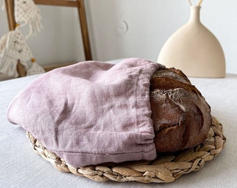 Puede incluir: Una hogaza de pan envuelta en una bolsa de pan de lino rosa polvoriento, sentada sobre un mantel individual de mimbre tejido.