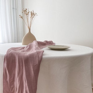 Peut inclure: Une nappe en lin blanc avec un chemin de table en lin rose et un petit vase avec des fleurs séchées sur une table ronde en bois.