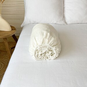 Puede incluir: Una sábana bajera de lino blanco enrollada en una cama blanca. La sábana está hecha de un tejido suave y natural, ideal para una noche de sueño confortable.