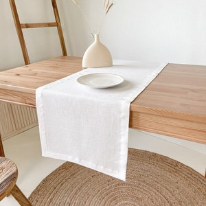 May include: White linen table runner on a wooden table with a white ceramic vase and dried wheat stalks. A white plate is on the table runner. The table is set on a woven rug.
