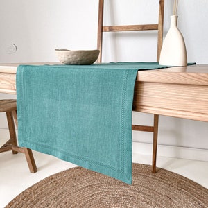 Könnte beinhalten: Ein tealfarbener Tischläufer aus Leinen mit einem gestickten Rand liegt auf einem Holztisch mit einem braunen Jute-Teppich darunter. Eine Holzleiter lehnt an einer weißen Wand hinter dem Tisch.