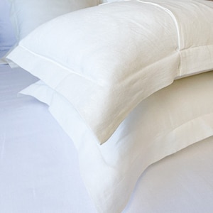 Pode incluir: Duas fronhas de linho branco com uma textura sutil e um design simples e limpo. As fronhas estão dobradas e empilhadas em uma cama branca.