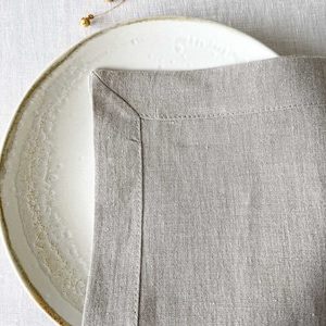 Könnte beinhalten: Nahaufnahme einer hellgrauen Leinenserviette mit einem weißen Teller darunter. Die Serviette hat einen genähten Rand und eine dezente Textur.