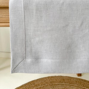 Puede incluir: Un camino de mesa de lino gris claro con una textura sutil y un borde cosido. El camino de mesa está drapeado sobre una mesa de madera.