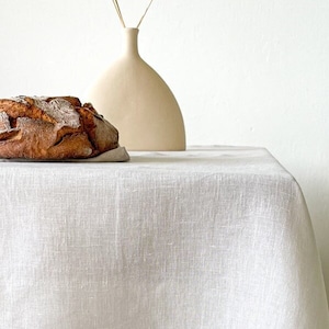 May include: White linen tablecloth with a stitched border, draped over a wooden table with a brown woven rug underneath. A loaf of bread sits on a linen napkin on the table, next to a white ceramic vase with dried grass.