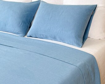 Puede incluir: Un juego de cama de lino azul con dos fundas de almohada y una sábana plana. La ropa de cama está en una cama con almohadas blancas y una alfombra tejida de color marrón claro.