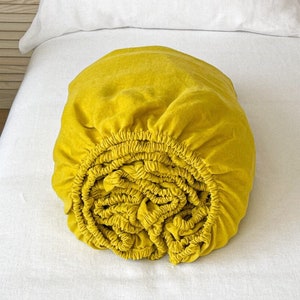 Pode incluir: Um lençol de elástico amarelo enrolado em uma cama branca. O lençol é feito de linho e tem um visual natural e relaxado.