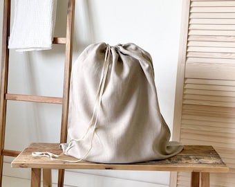 May include: A beige linen drawstring laundry bag rests on a rustic wooden bench. A wooden ladder with a white towel leans against the wall. The scene is illuminated by natural light, creating a minimalist aesthetic.