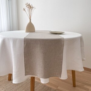 Peut inclure: Une nappe ronde blanche avec un chemin de table en lin beige sur une table en bois. La table est dressée avec une petite assiette et un vase de fleurs séchées.
