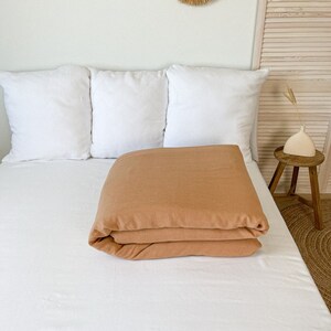 May include: A folded terracotta-colored linen duvet cover on a white bed with three white pillows. The bed is against a light beige wall with a woven wall hanging above it.