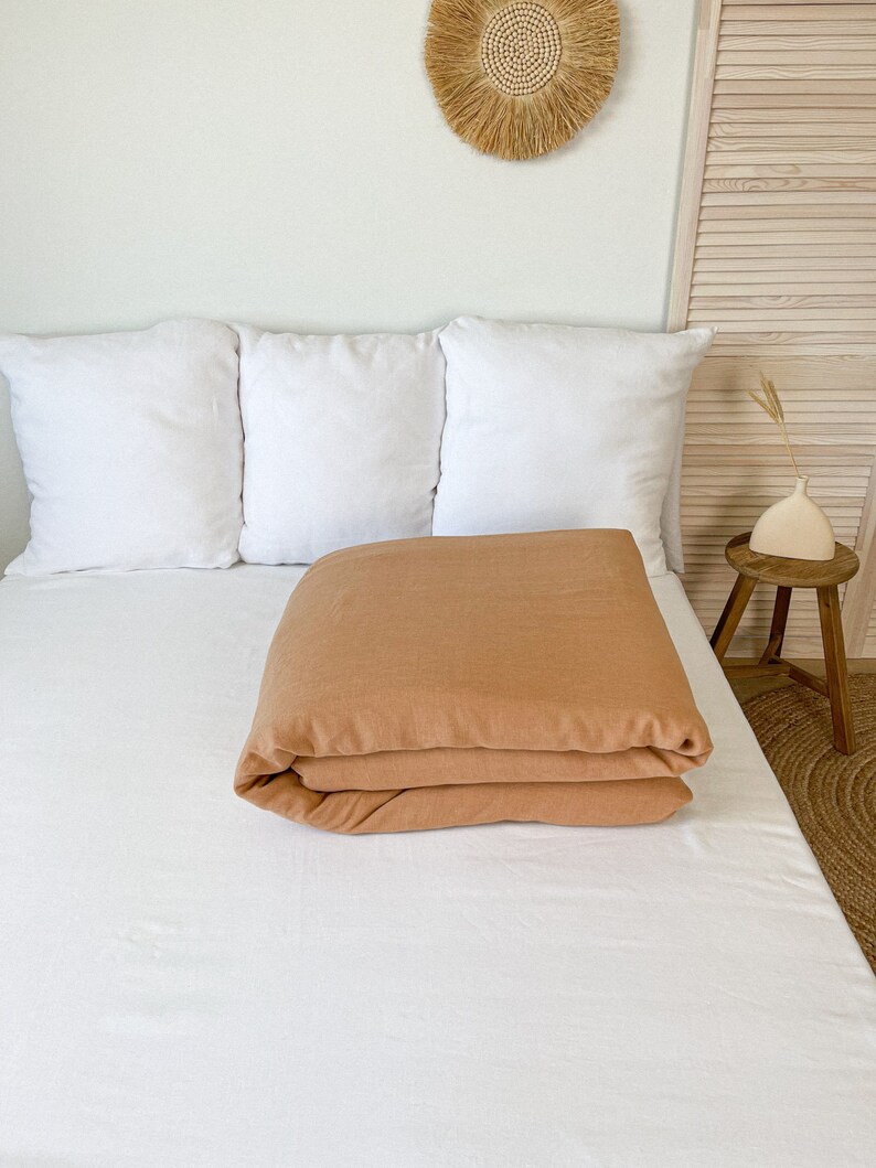 May include: A folded terracotta-colored linen duvet cover on a white bed with three white pillows. The bed is against a light beige wall with a woven wall hanging above it.