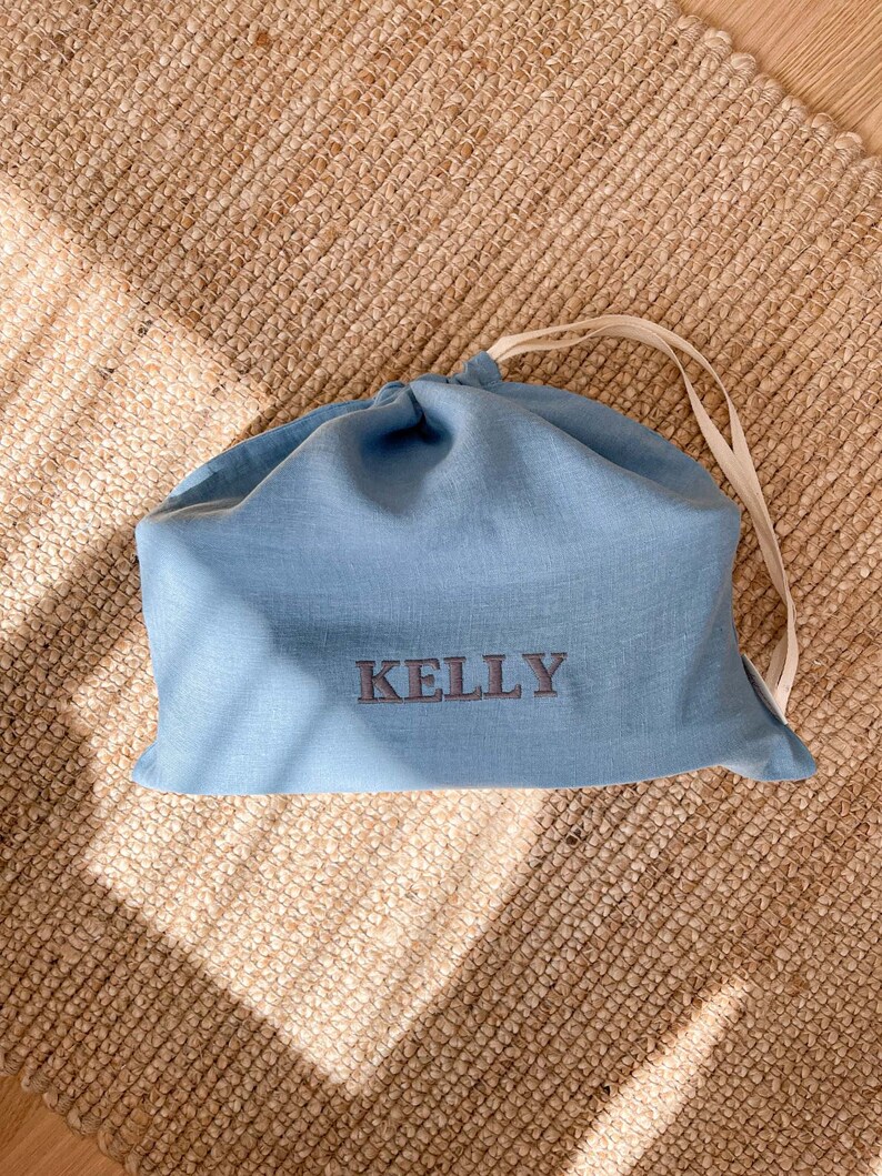 May include: A light blue linen drawstring bag with the name "Kelly" embroidered in dark gray thread.