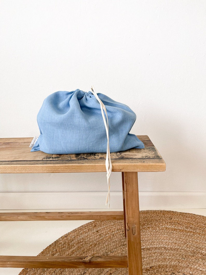 May include: A light blue linen drawstring bag sits on a wooden bench. The bag has a white drawstring and is made of a soft, natural fabric.