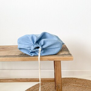 May include: A light blue linen drawstring bag sits on a wooden bench. The bag has a white drawstring and is empty.