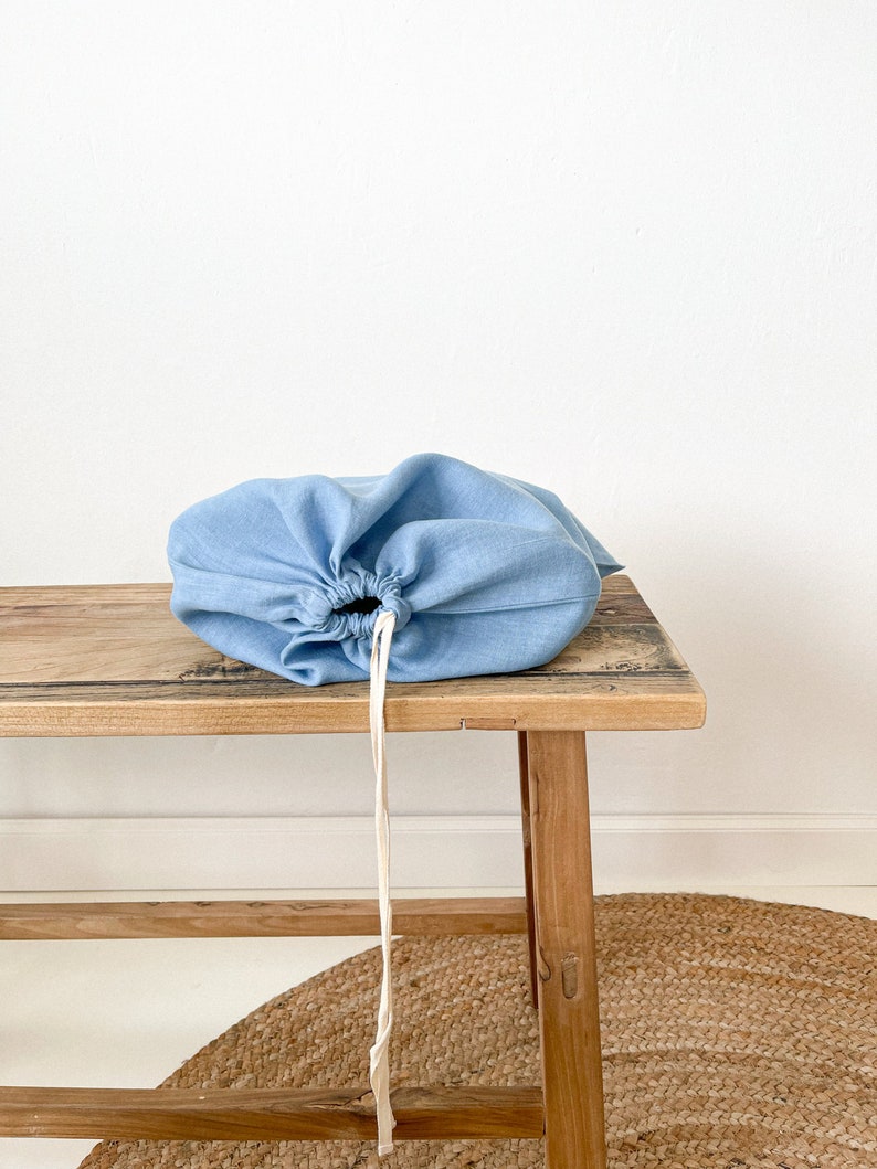 May include: A light blue linen drawstring bag sits on a wooden bench. The bag has a white drawstring and is empty.