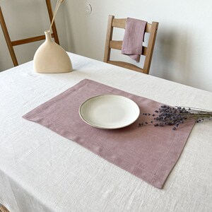 Puede incluir: Un mantel individual de lino rosa polvoriento con un plato de cerámica blanco en la parte superior. El mantel individual está sobre un mantel de lino blanco. Hay un ramo de flores de lavanda secas sobre la mesa.