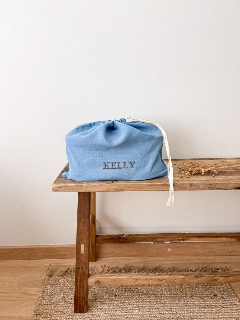 May include: A light blue linen storage bag with the name "KELLY" embroidered on the front. The bag is sitting on a wooden bench with a natural finish.