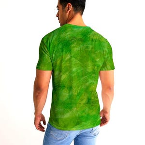 May include: A green t-shirt with a textured, watercolor-like design. The shirt is short-sleeved and has a crew neck. The model is wearing blue jeans.