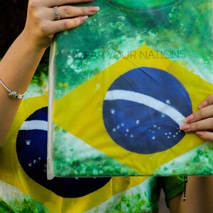 May include: A green, yellow, and blue t-shirt featuring the Brazilian flag design. The shirt has the text "WEAR YOUR NATIONS" printed on it. The shirt is held up by a person.