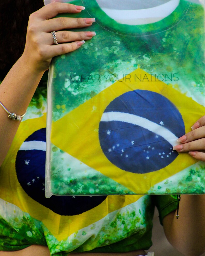May include: A green, yellow, and blue t-shirt featuring the Brazilian flag design. The shirt has the text "WEAR YOUR NATIONS" printed on it. The shirt is held up by a person.