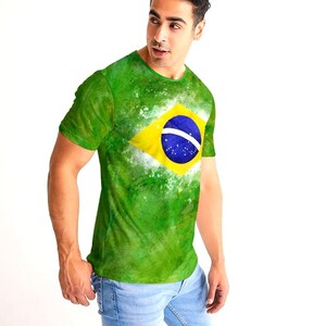 May include: Green t-shirt featuring the Brazilian flag. The shirt has a yellow diamond with a blue circle and white stars in the center. The background is a watercolor-style green with white accents. The person is wearing light blue jeans.