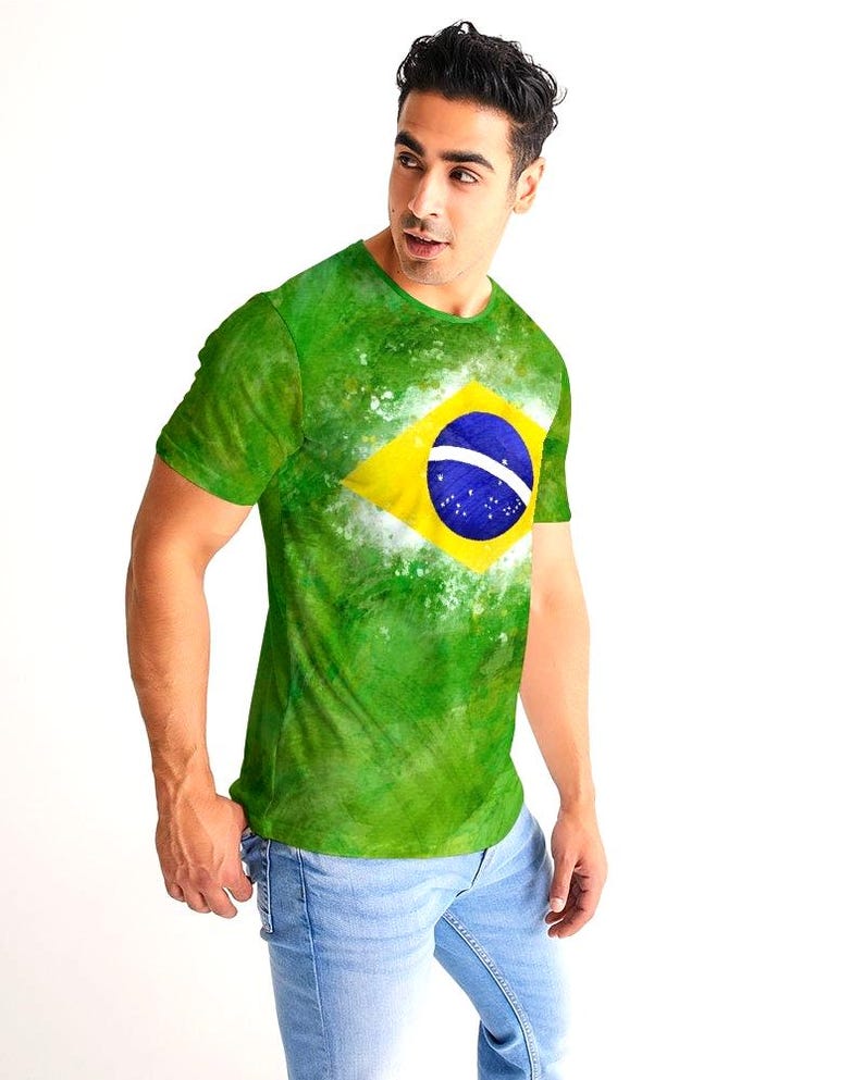 May include: Green t-shirt featuring the Brazilian flag. The shirt has a yellow diamond with a blue circle and white stars in the center. The background is a watercolor-style green with white accents. The person is wearing light blue jeans.