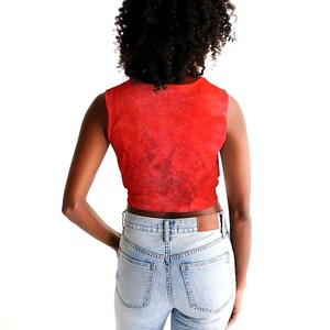 May include: A sleeveless red crop top with a textured pattern. The top is paired with light blue denim jeans. The model is facing away from the camera, with hands in the pockets of the jeans.