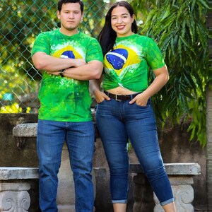 May include: Two people wearing green tie-dye t-shirts with the Brazilian flag design. They are also wearing blue jeans. The background includes a green fence and foliage.