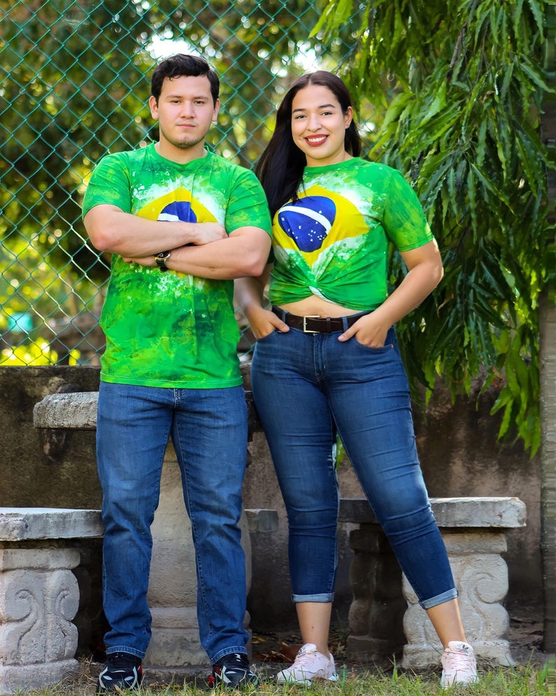 May include: Two people wearing green tie-dye t-shirts with the Brazilian flag design. They are also wearing blue jeans. The background includes a green fence and foliage.