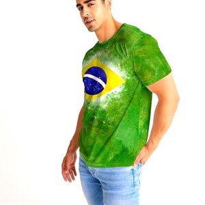 May include: Green t-shirt featuring the Brazilian flag design. The shirt has a yellow diamond with a blue circle and white stripe. The shirt is worn with light blue jeans. The t-shirt has a watercolor effect.
