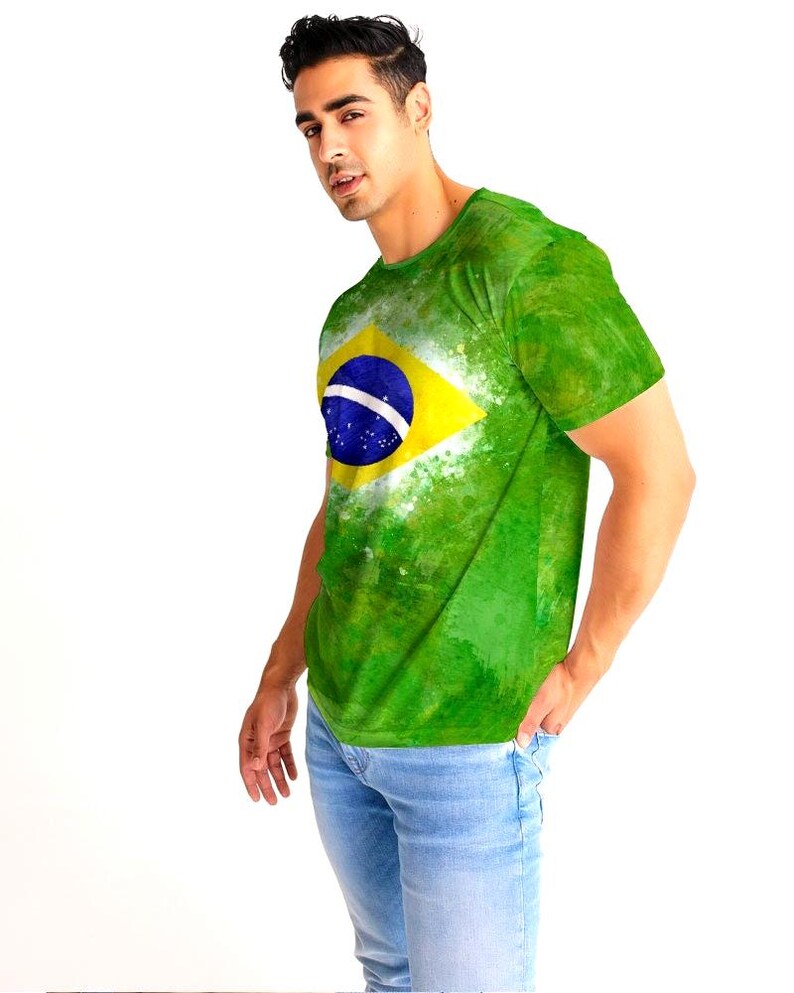 May include: Green t-shirt featuring the Brazilian flag design. The shirt has a yellow diamond with a blue circle and white stripe. The shirt is worn with light blue jeans. The t-shirt has a watercolor effect.