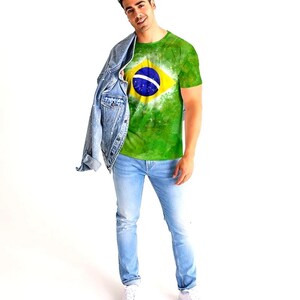 May include: A green t-shirt featuring the Brazilian flag, with a yellow diamond and a blue circle containing a white band. The shirt is worn with light blue jeans, a denim jacket, and white sneakers. The person is smiling.