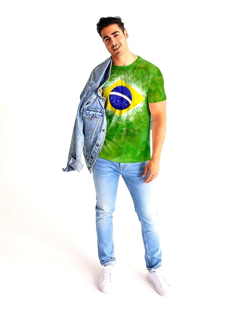 May include: A green t-shirt featuring the Brazilian flag, with a yellow diamond and a blue circle containing a white band. The shirt is worn with light blue jeans, a denim jacket, and white sneakers. The person is smiling.