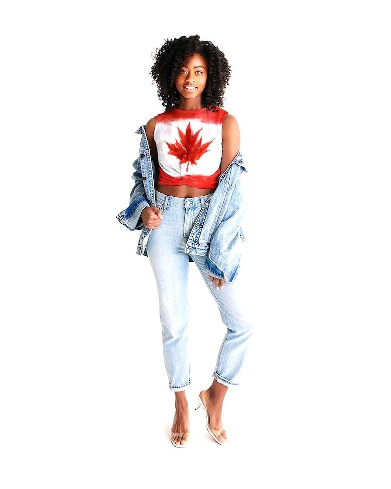 May include: A red and white crop top with a large red maple leaf design. The top is paired with light blue jeans and a denim jacket. The person is wearing clear heeled sandals.