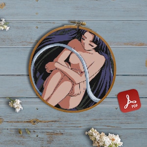 May include: A cross-stitch pattern of a woman with long dark hair, sitting with her arms wrapped around her legs. The pattern is in a wooden embroidery hoop on a blue wooden background.