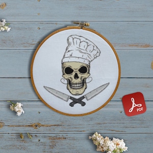 May include: A white embroidery hoop with a black and white skull wearing a chef's hat and holding two crossed knives. The hoop is on a light blue wooden surface with white flowers and colorful embroidery floss.